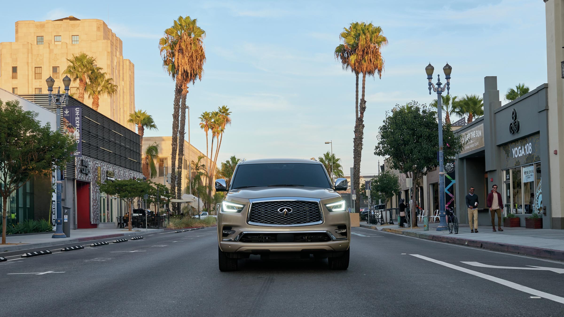 A beige 2022 INFINITI QX80 SUV driving down a street with palm trees and shops.